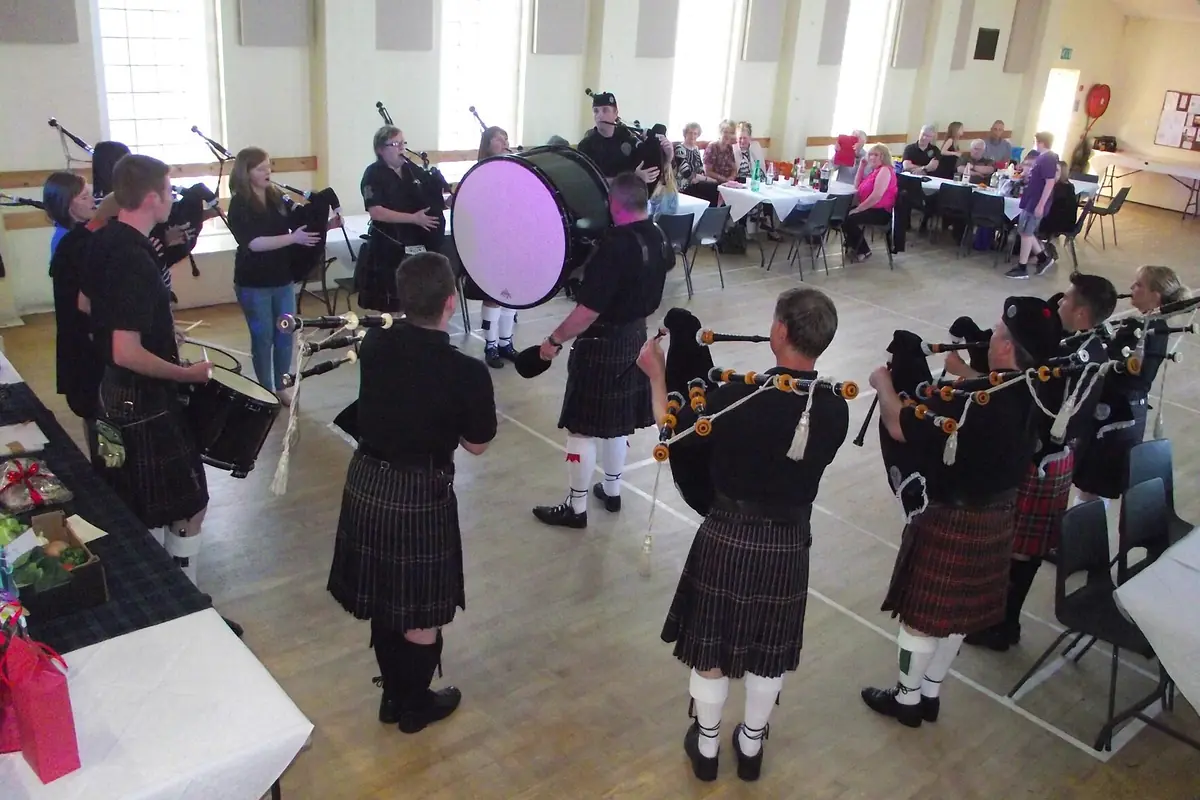 A pipe band in kilts plays in a village hall with a wooden floor while guests sit at tables behind them