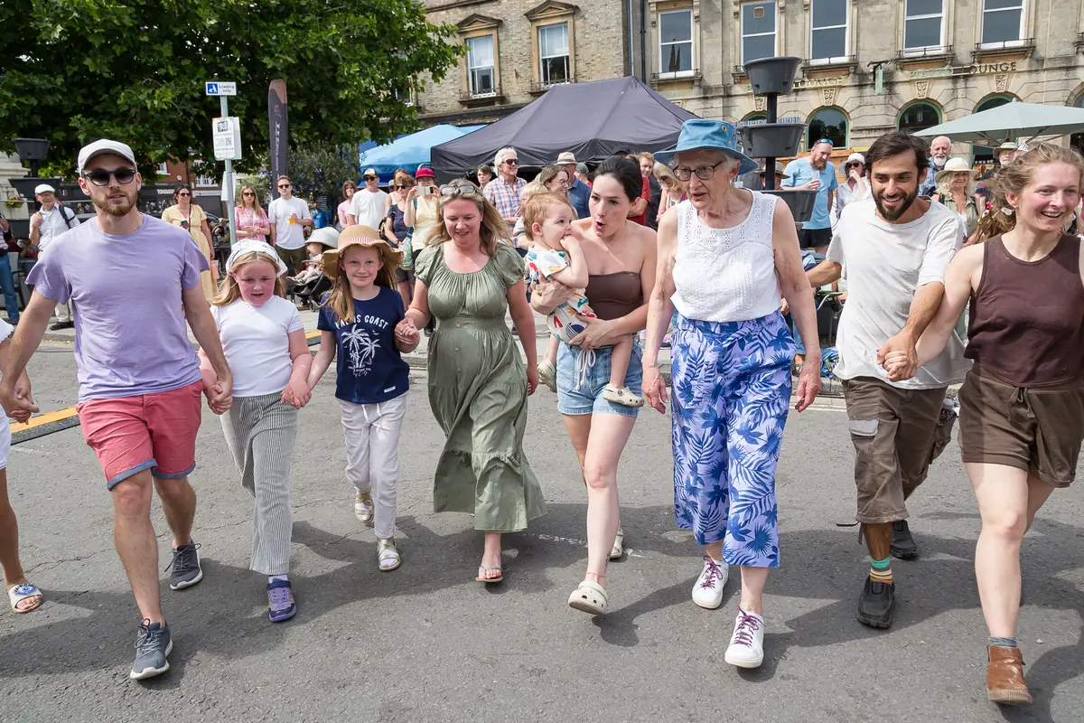 A line of ceilidh dancers holding hands and walking along a closed street in front of a crowd, with a stone building behind