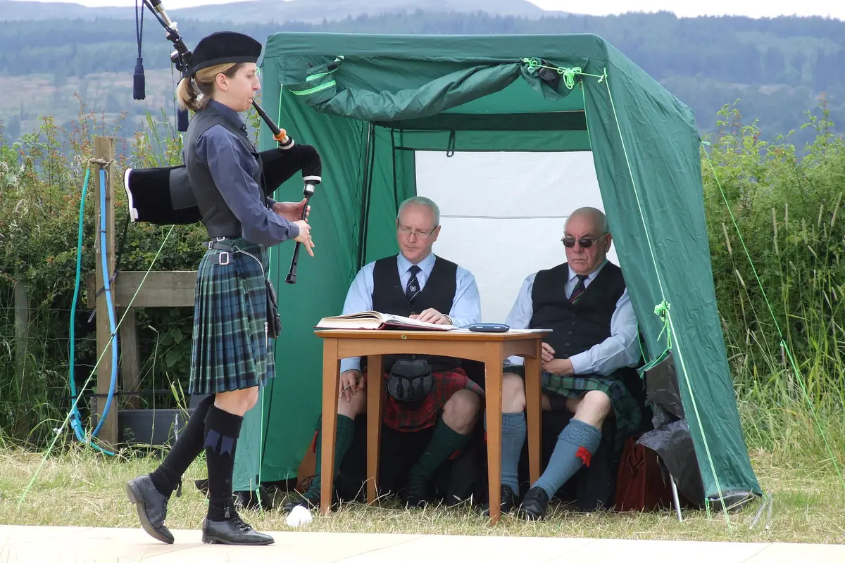 A piper in a bonnet and tartan kilt walks past a green tent where two judges sit at a table, with hills behind