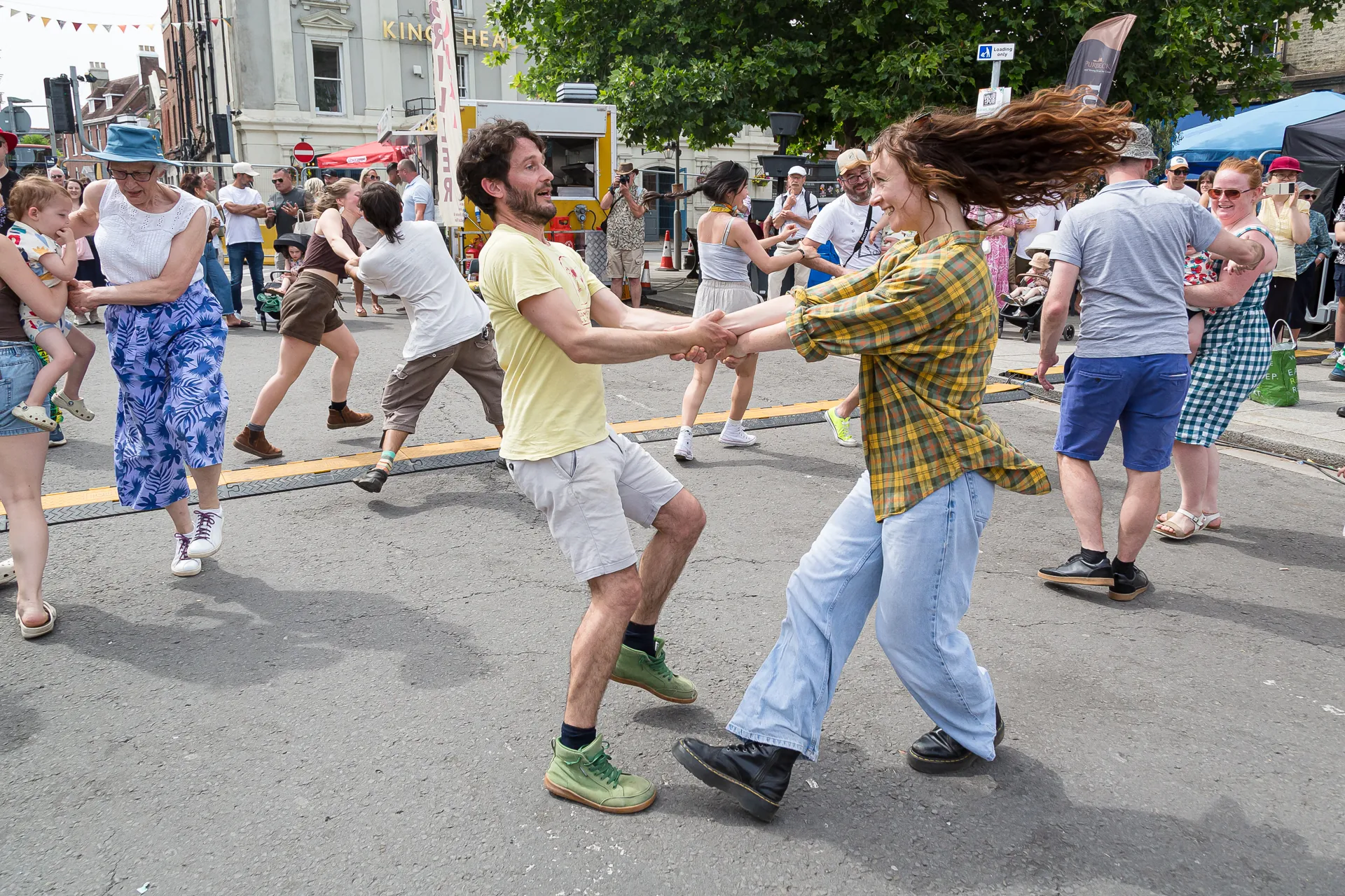 Two dancers swing each other by the hands in a ceilidh set on a closed street at a folk festival in Dorset, England, with other dancers and onlookers around them